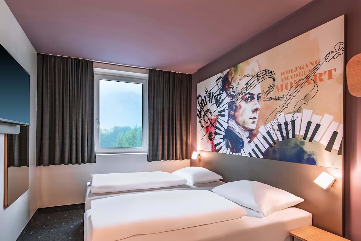 A hotel room with two beds, a large window with curtains, a mural on the wall, and a small wooden table.