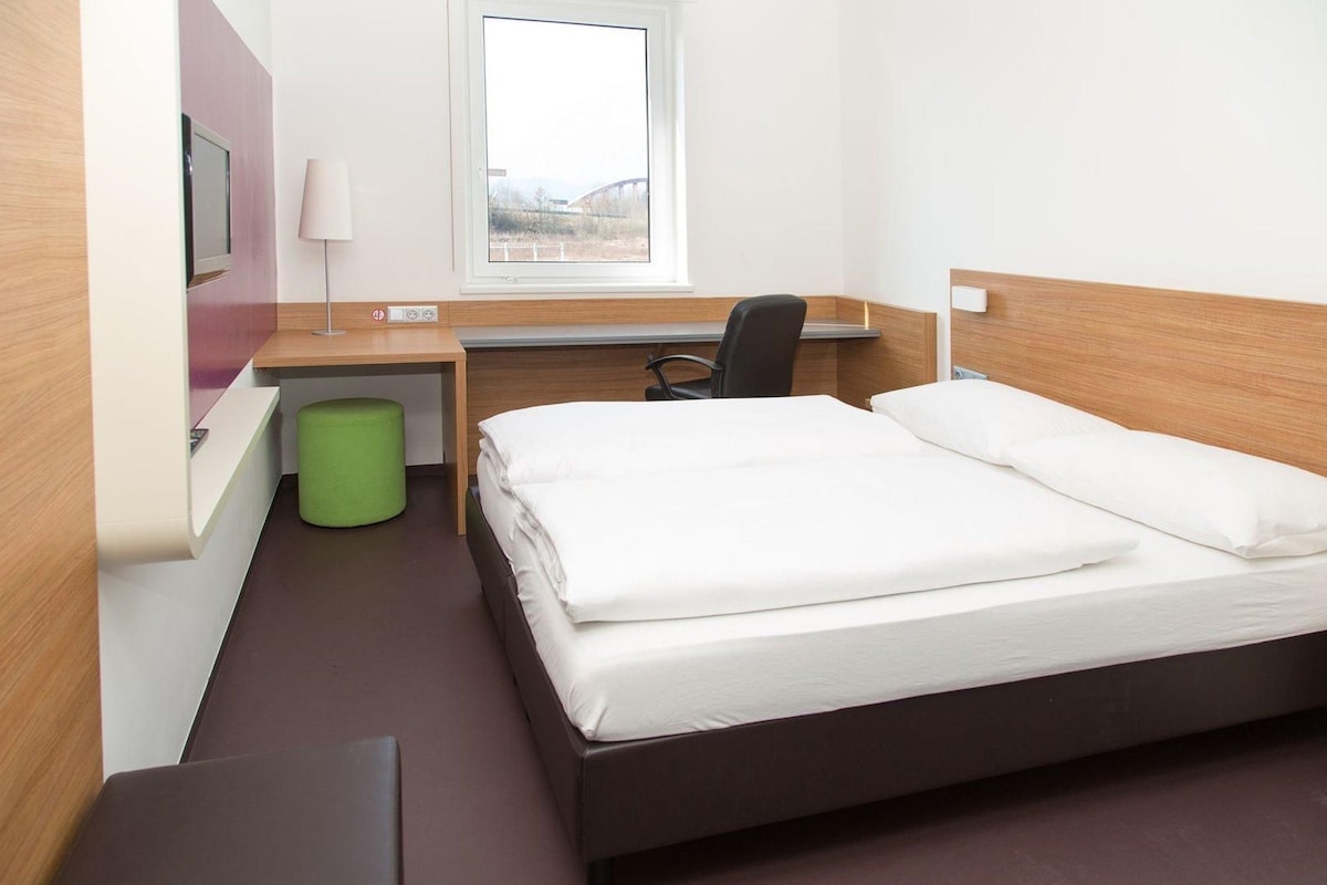 Une chambre d’hôtel avec un lit, un bureau, une chaise, une poubelle verte et une fenêtre donnant sur un pont.