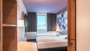 Une chambre d’hôtel avec un lit, un bureau, une chaise, une fenêtre avec des rideaux et un mur orné d’une affiche.