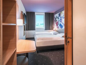 A hotel room with a bed, a desk, a chair, a window with curtains, and a wall with a poster.
