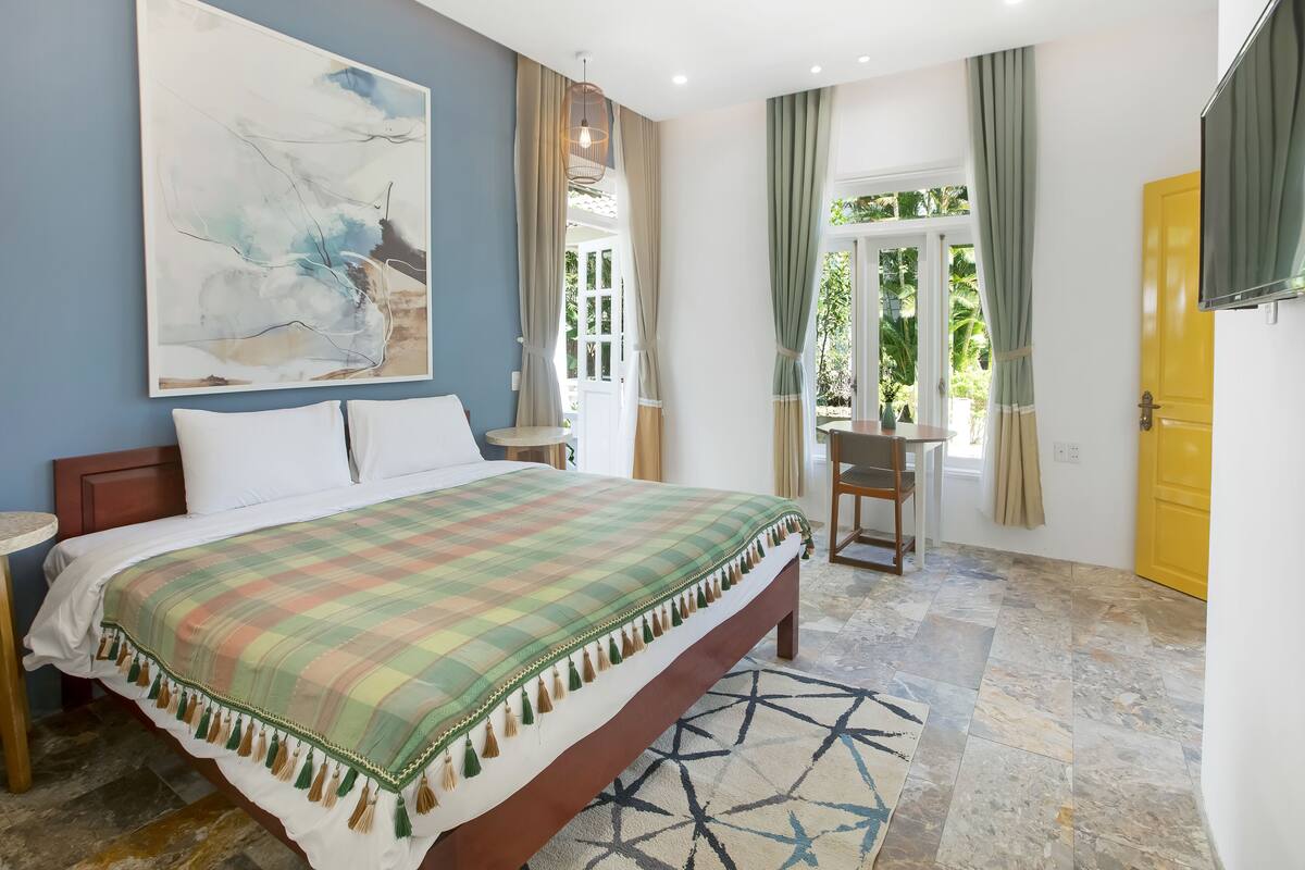 Une chambre à coucher avec un lit, un grand tableau abstrait, un bureau et une fenêtre avec des rideaux.