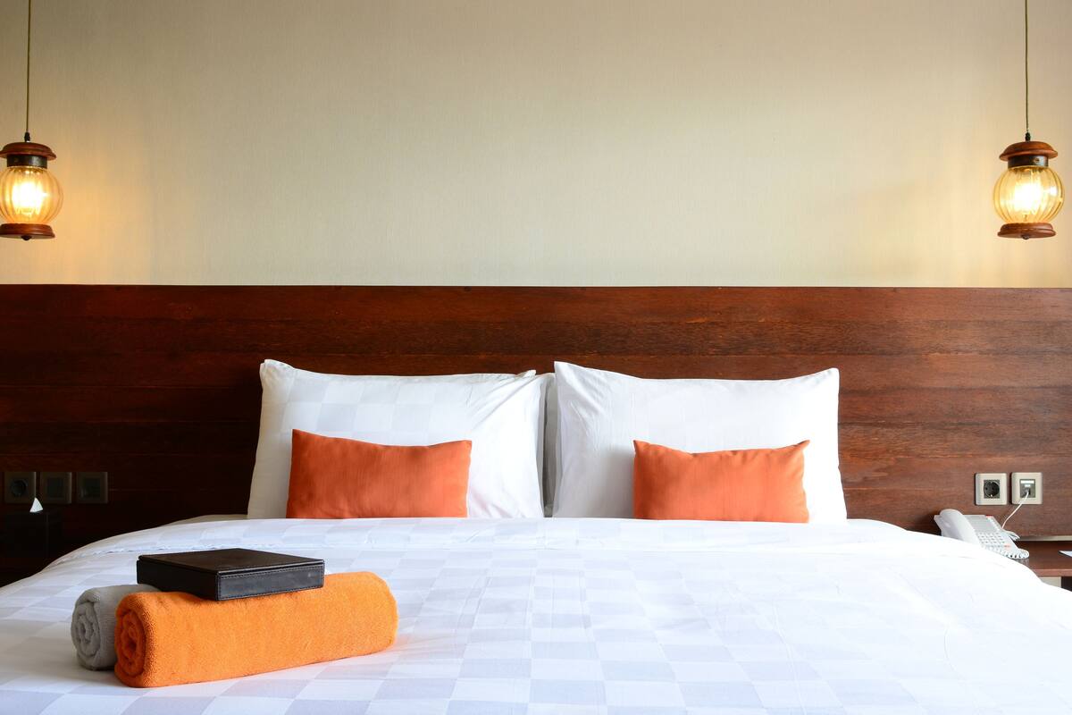 A neatly made bed with white and orange pillows, a rolled-up towel, and a black object on the bed. There are two wall-mounted light fixtures above the bed.