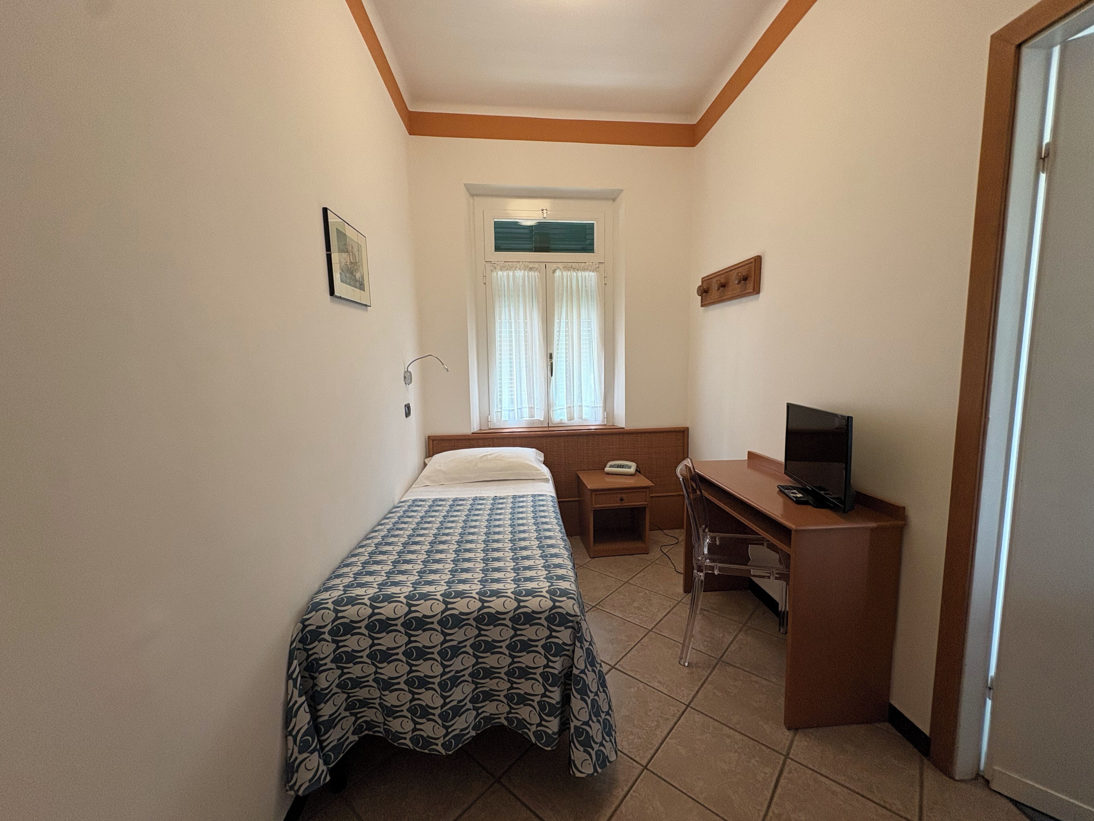 Una habitación pequeña con cama, escritorio con computadora, ventana con cortinas y puerta.