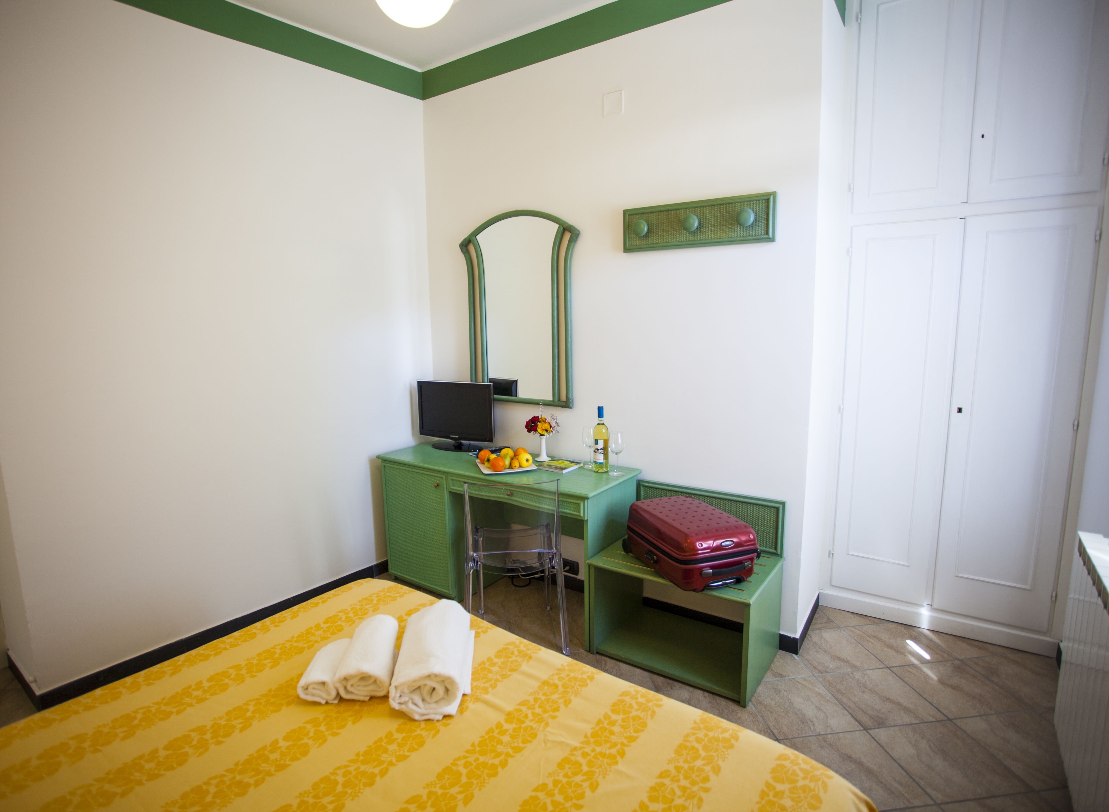 A hotel room with a bed, a desk with a computer, a mirror, a small table with a vase of flowers, a bottle of wine, and a suitcase.