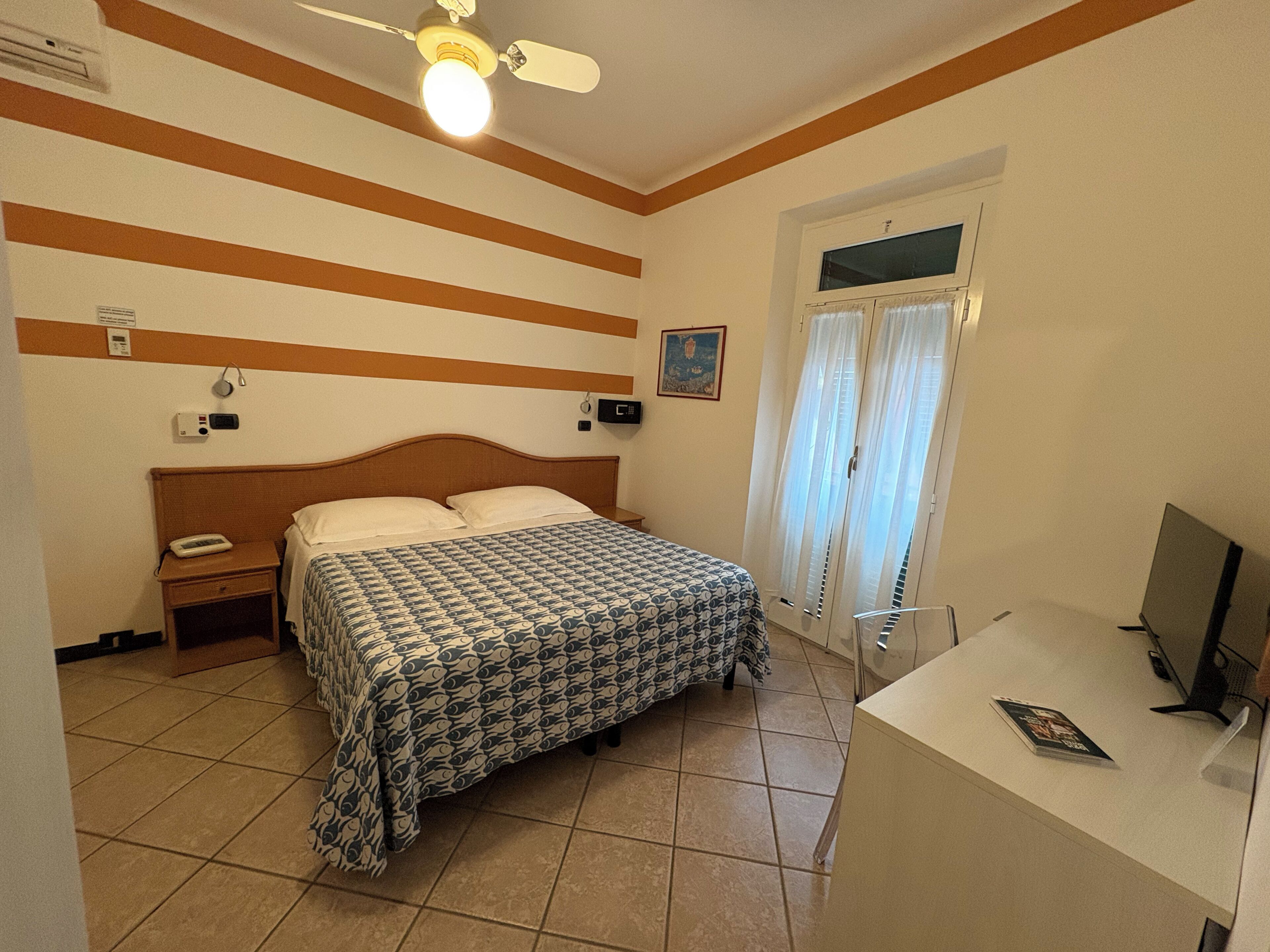 A hotel room with a bed, a desk, a chair, a ceiling fan, a window with curtains, and a wall-mounted air conditioner.