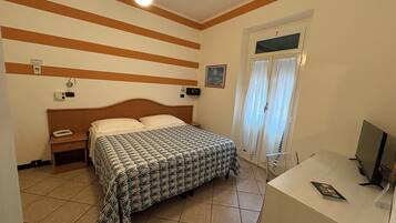 A hotel room with a bed, a desk, a chair, a ceiling fan, a window with curtains, and a wall-mounted air conditioner.