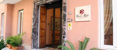 Entrada de un hotel con puerta enmarcada en mármol, un letrero que dice 'Hotel delle Rose' y una estación de recepción.