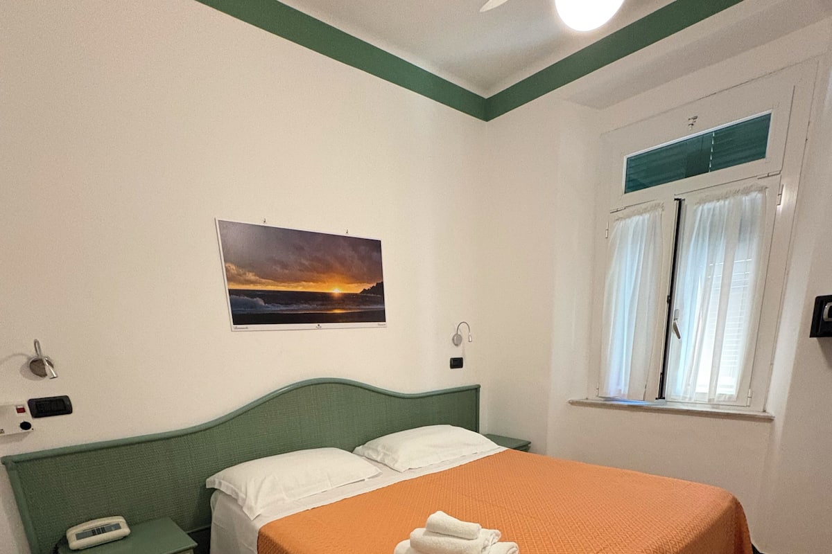 A bedroom with a bed, a nightstand, a bedside table, a window with curtains, a wall-mounted picture, and a ceiling fan.