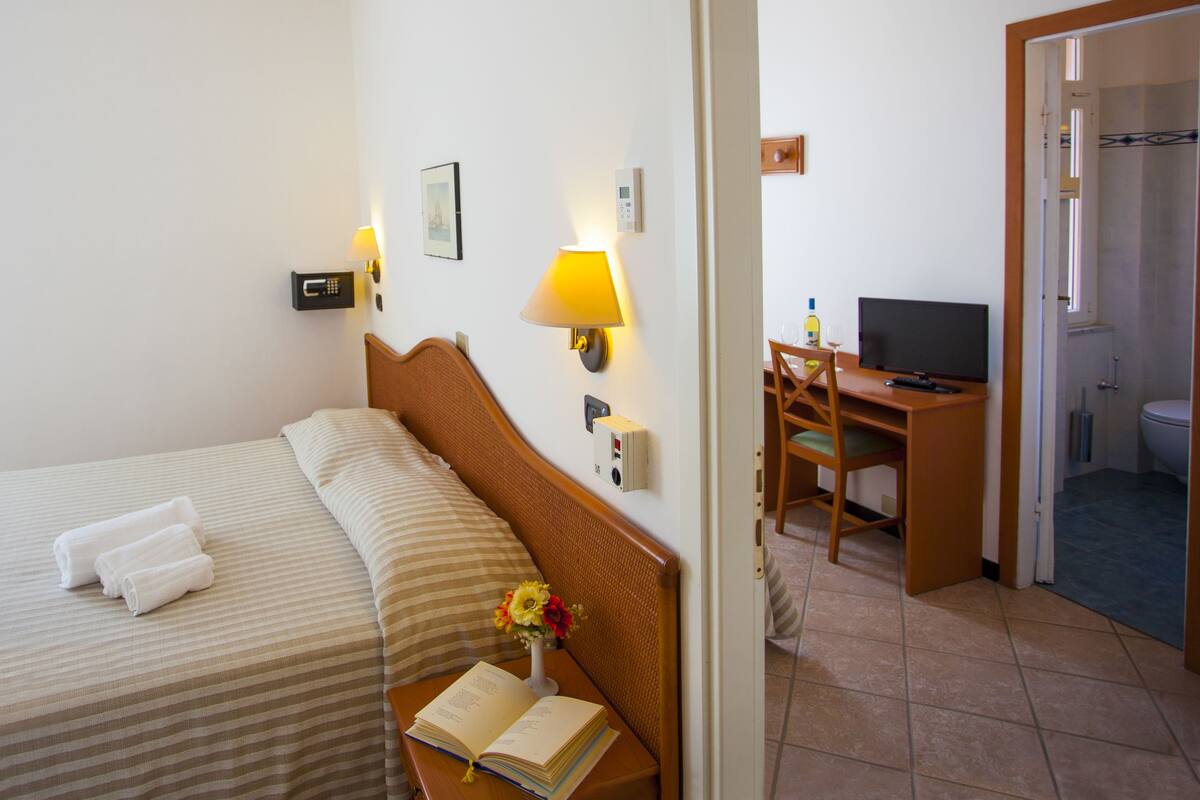 A hotel room with a bed, a desk with a television, a chair, a lamp, and a small vase with flowers.