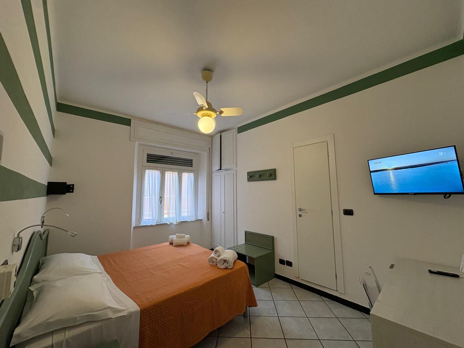Quarto de hotel com uma cama, televisão na parede, janela com cortinas, porta e ventoinha no teto.
