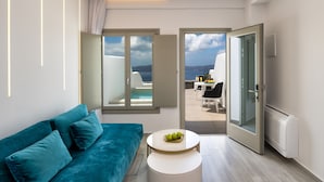 Un salon moderne avec un canapé de couleur turquoise, une table basse ronde et blanche ornée de fruits, et une vue sur la mer par des portes ouvertes.