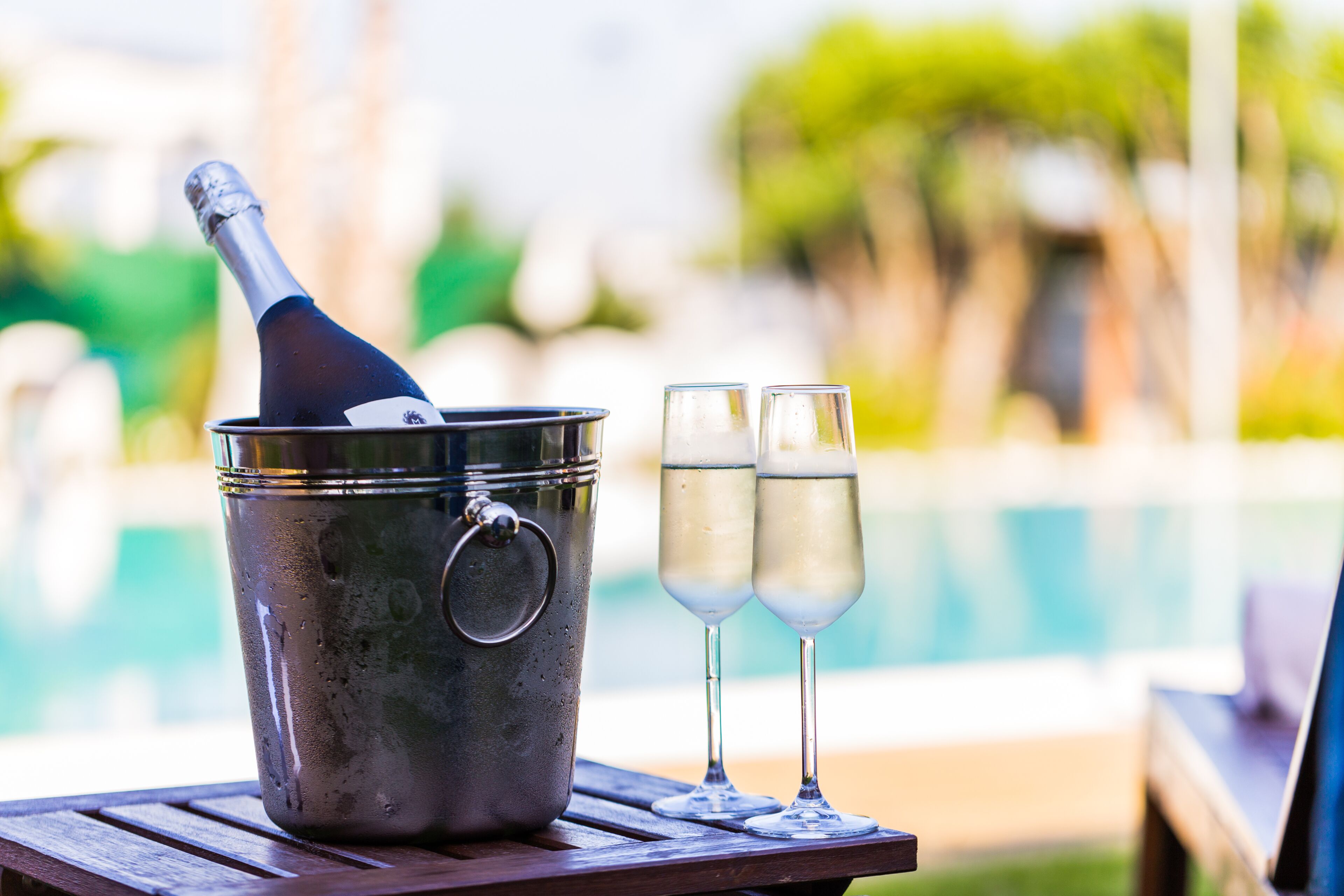 A metal bucket with a bottle opener and two glasses of champagne on a wooden table by a pool.