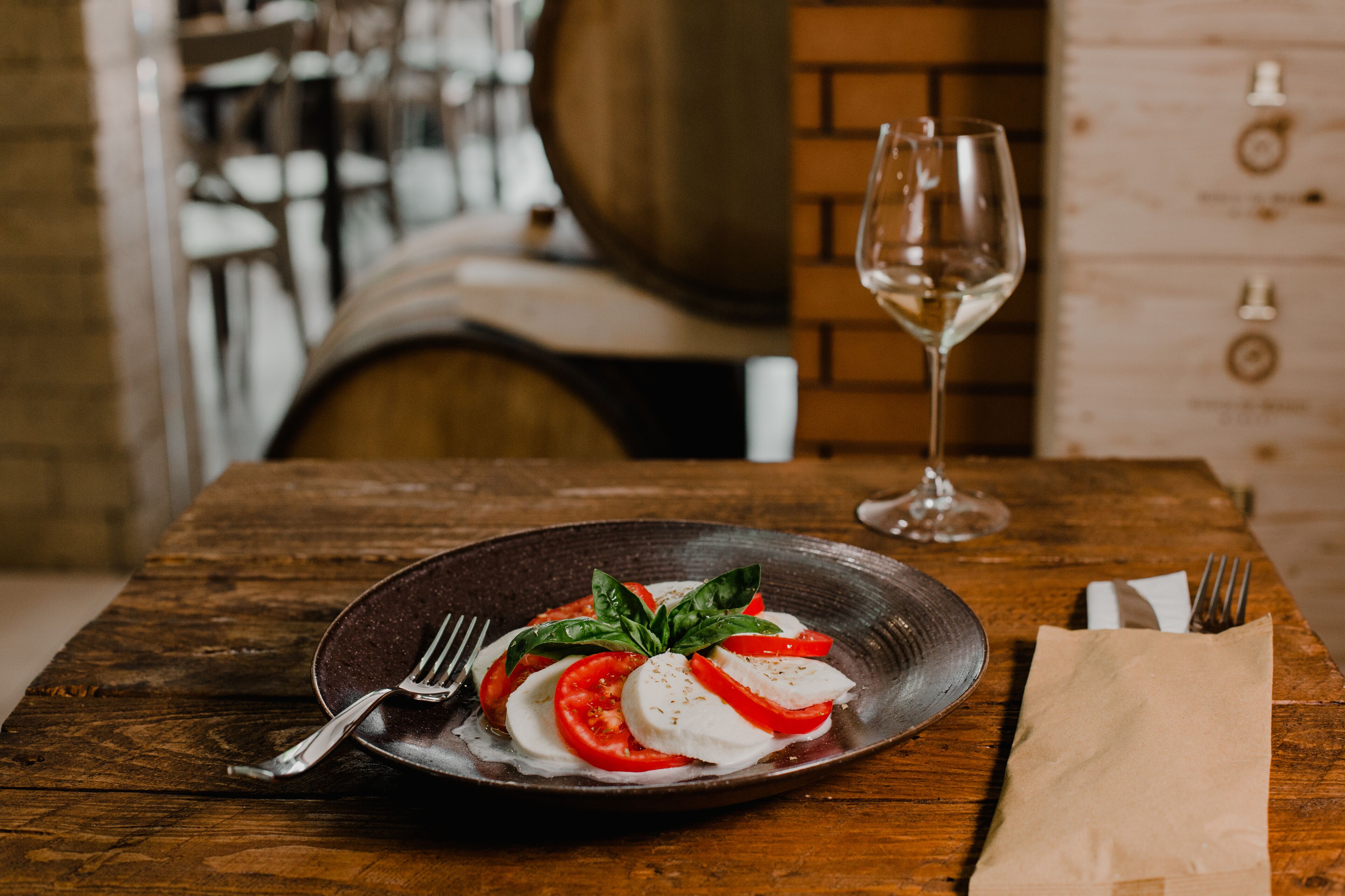 Una mesa de madera con una ensalada caprese, un tenedor, una servilleta y una copa de vino.