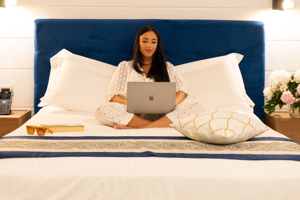 Eine Person sitzt auf einem Bett mit einem Laptop, einem Handy und einem Buch auf einem Nachttisch.