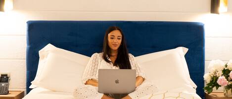 Eine Person sitzt auf einem Bett mit einem Laptop, einem Handy und einem Buch auf einem Nachttisch.