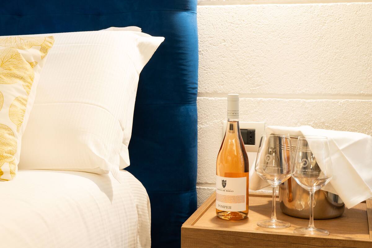 Ein ordentlich bezogenes Bett mit blauem Kopfteil, weißen Kissen und einem gelben Blattmuster-Kissen. Ein Nachttisch neben dem Bett enthält eine Weinflasche, ein Glas und einen Metallbehälter, möglicherweise für Wasser oder andere Getränke.