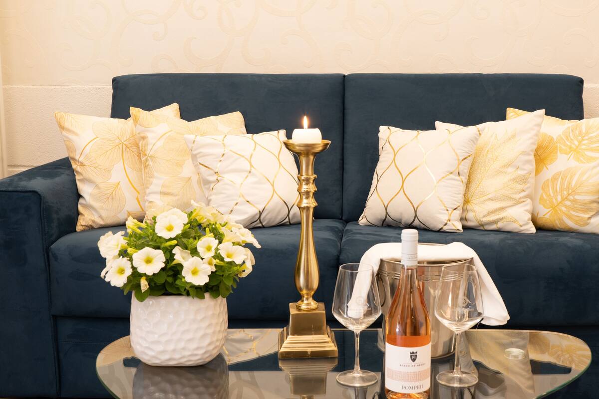 Ein dunkelblaues Sofa mit Kissen in Gold- und Weißmuster, ein kleiner Tisch mit einer Vase weißer Blumen, einer Kerze und zwei Weingläsern.