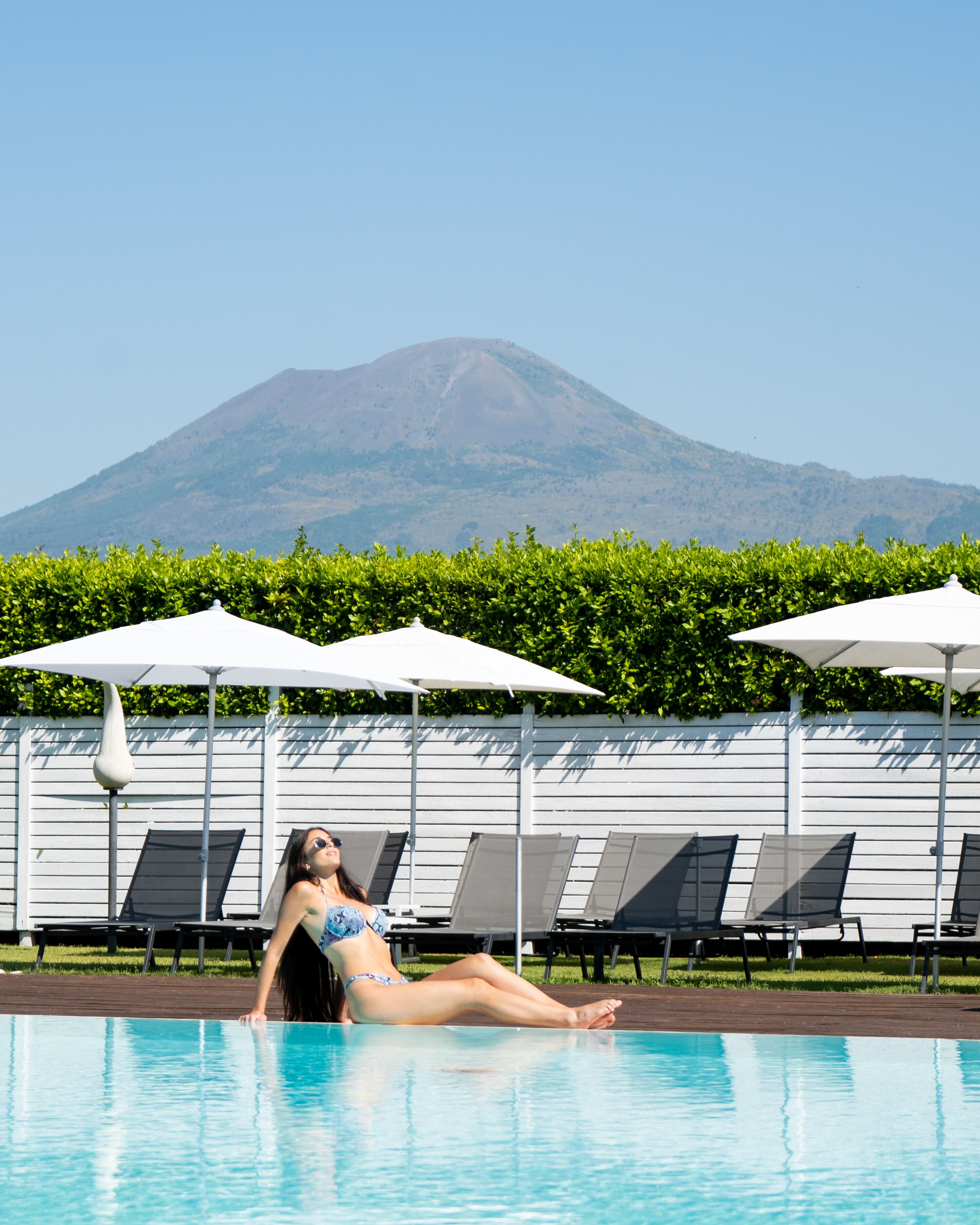 A pool area with lounge chairs and umbrellas, a woman relaxing in the water, and a mountain in the background.