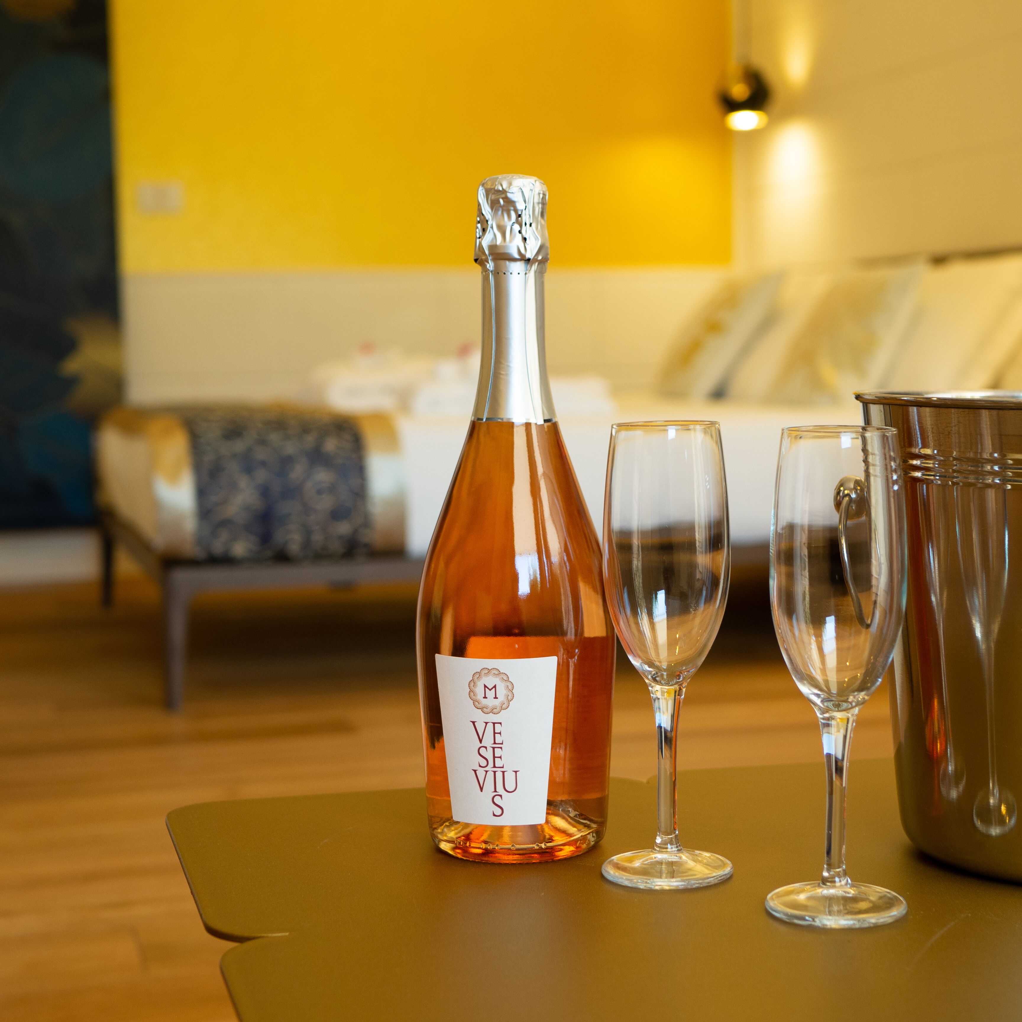 A bottle of Veuve's wine, two wine glasses, and a metal bucket on a table in a room with a bed and a yellow wall in the background.