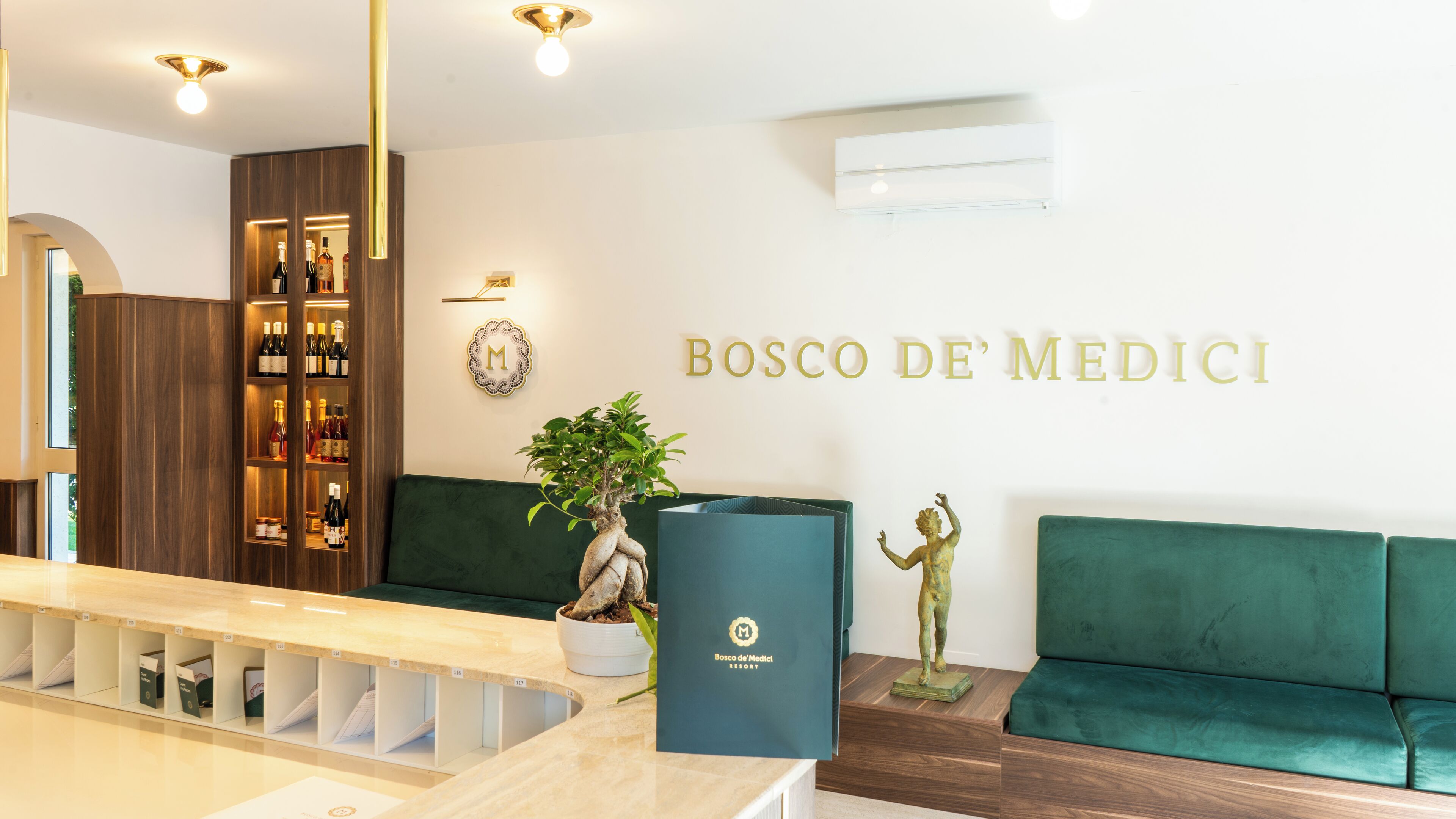 A reception area with a wooden reception desk, a green sofa, a potted plant, a statue, and a wall with the text "BOSCO DE MEDICI".