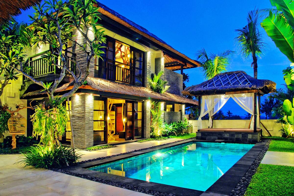 A two-story house with a swimming pool, surrounded by tropical plants and a thatched-roof gazebo.