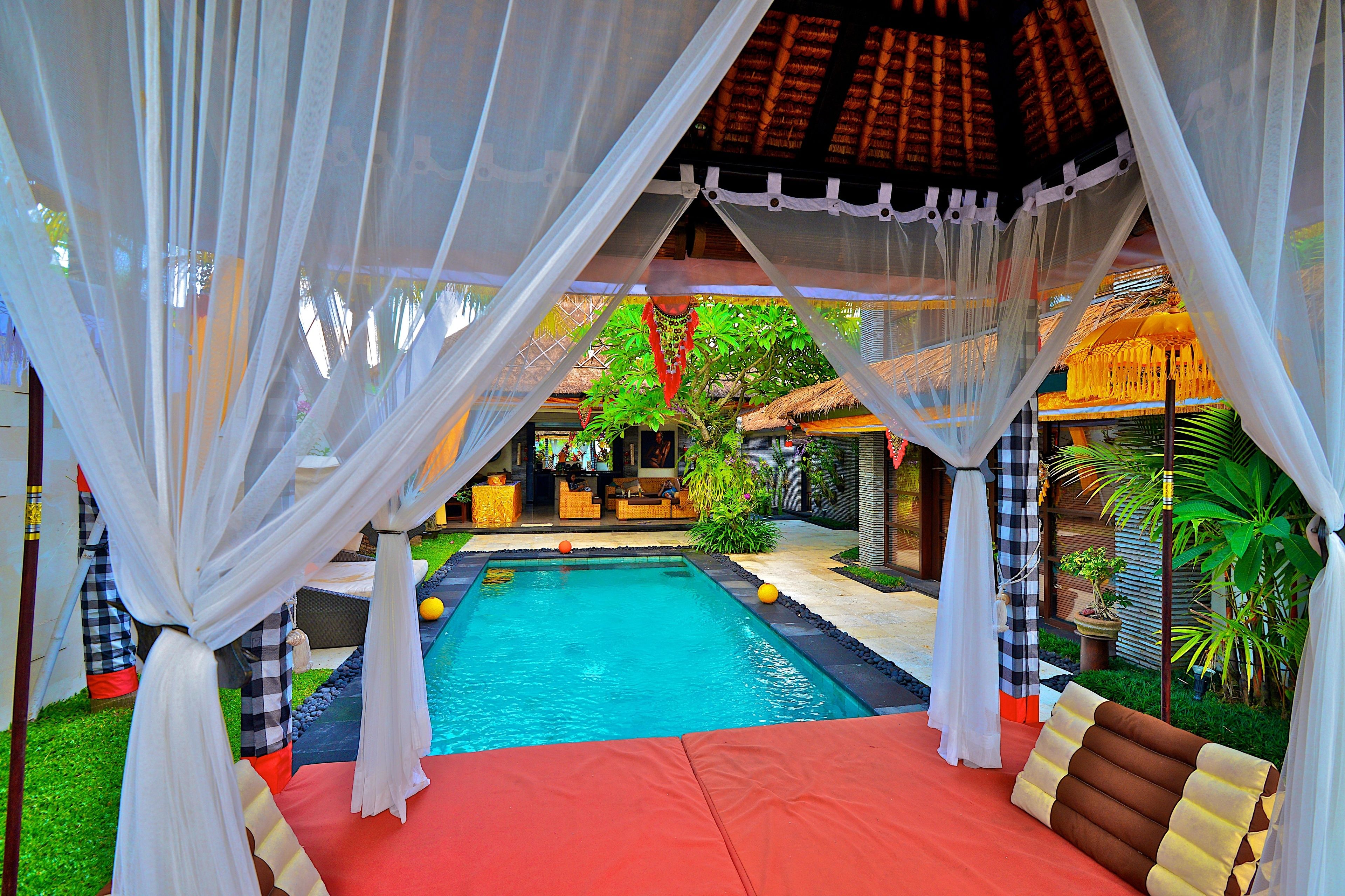 A pool area with a red deck, white canopy, and a thatched-roof structure in the background.