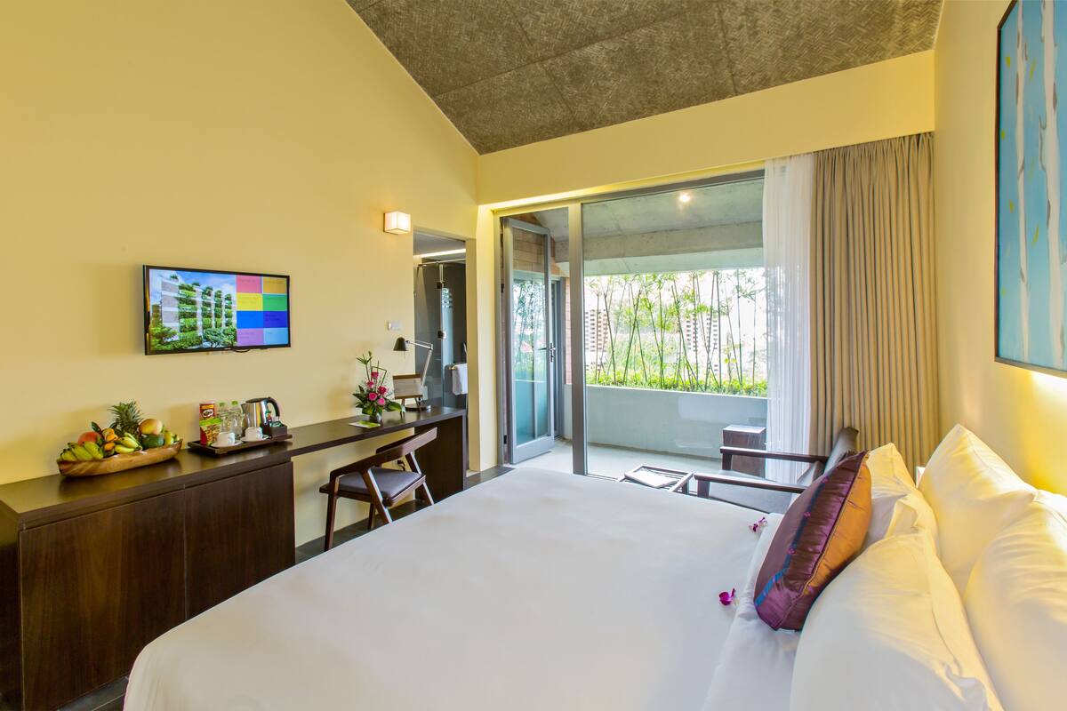 A modern hotel room with a large bed, a desk with a computer, a flat-screen TV, and a balcony with a view of greenery.