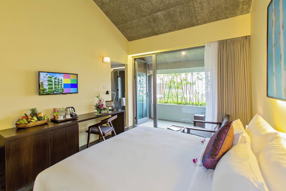 A modern hotel room with a large bed, a desk with a computer, a flat-screen TV, and a balcony with a view of greenery.