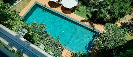 A rectangular swimming pool with a wooden deck, surrounded by lush greenery and palm trees.