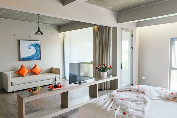 A modern hotel room with a sofa, a flat-screen TV, a dining table, and a bed with rose petals.