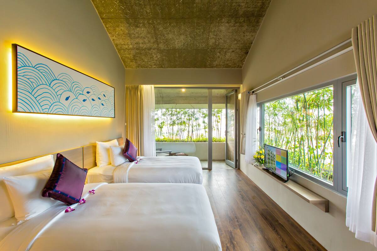 A hotel room with two beds, a large window with a view of greenery, and a painting on the wall.