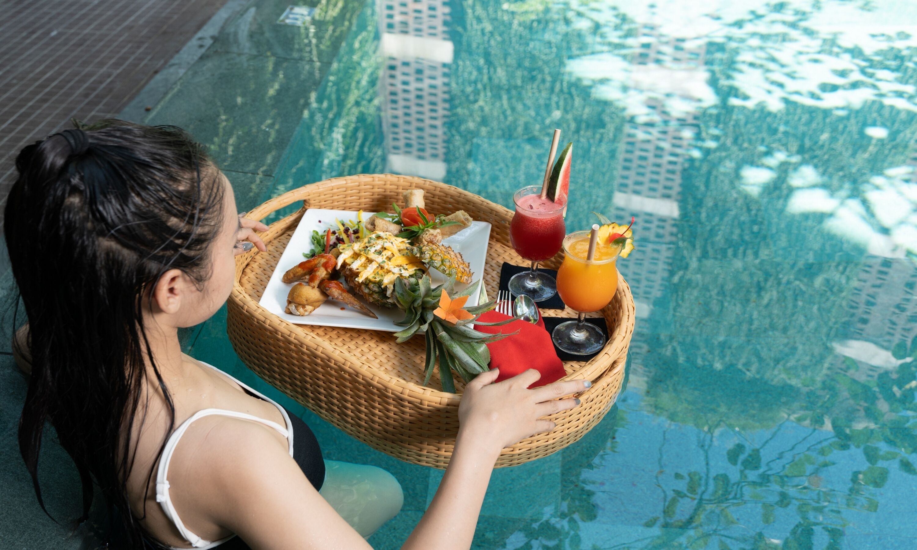 A person sitting by a poolside table with a meal and drinks.