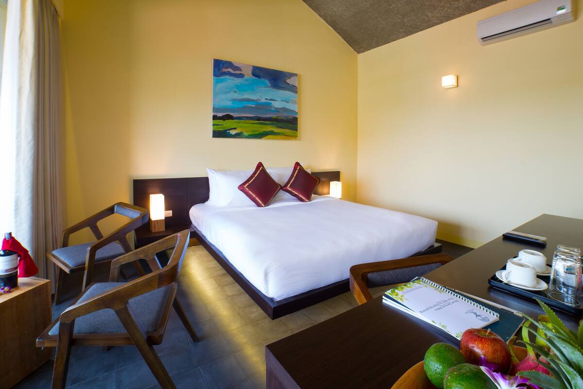 A hotel room with a bed, bedside tables, a desk, a chair, a painting on the wall, and a window with curtains.