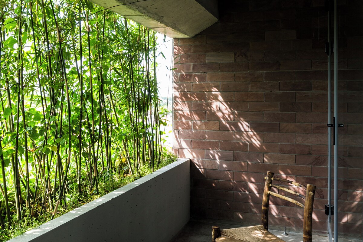 Una habitación con techo de concreto, ventana con vista a bambú, una silla de madera y una mesita.
