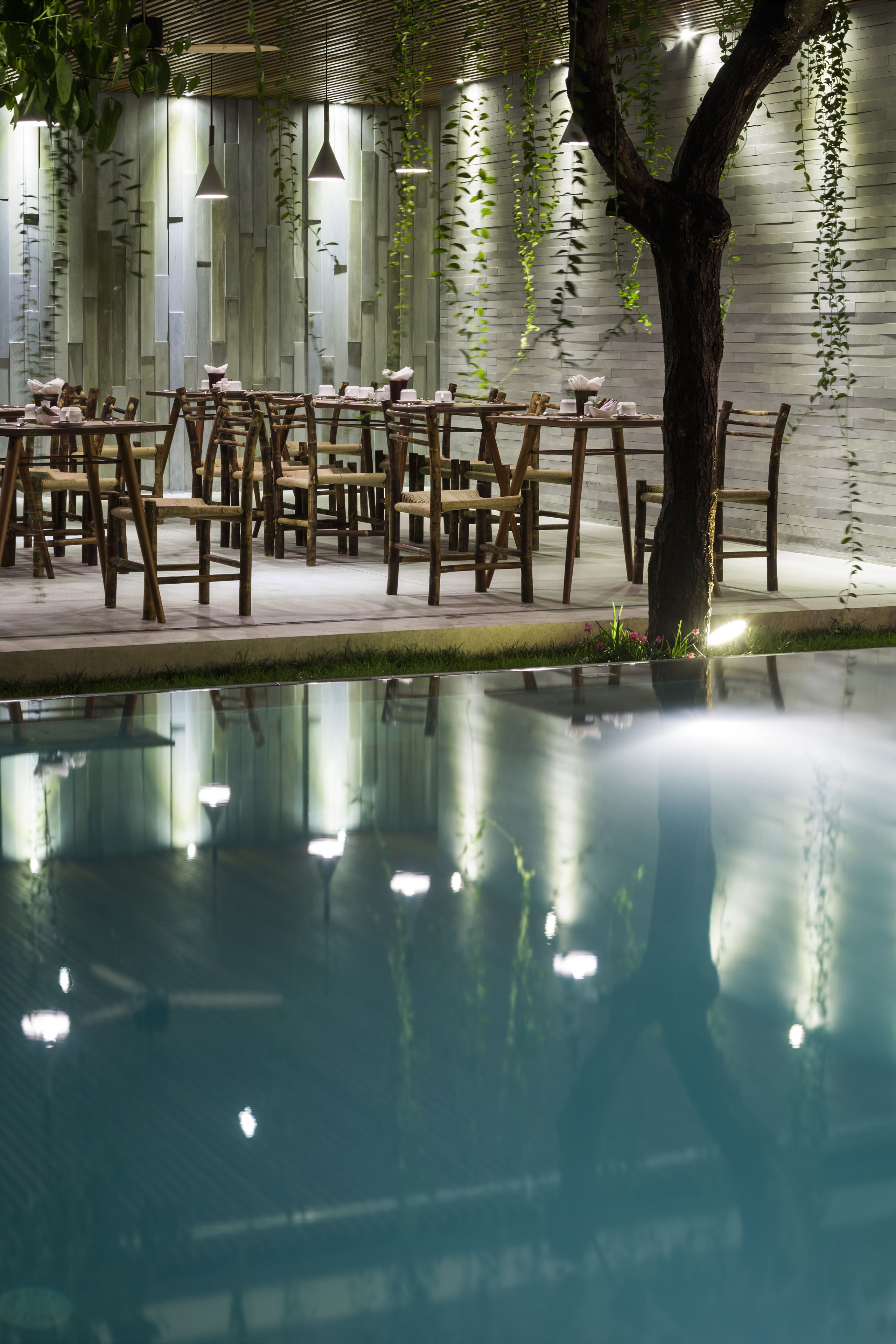 A serene indoor pool area with wooden tables and chairs, a tree, and hanging plants against a white brick wall.