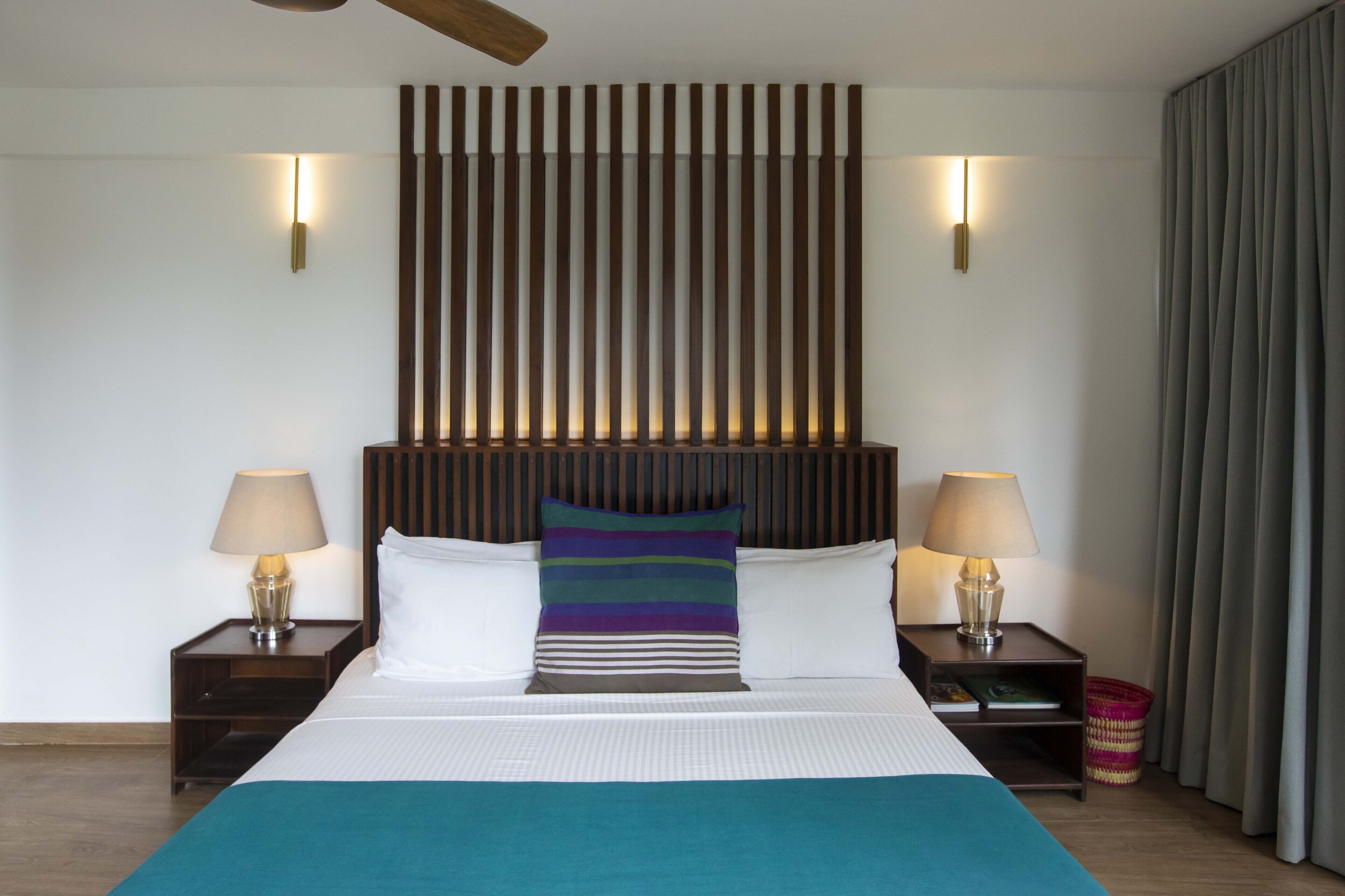 A modern hotel room with a large bed, two bedside tables with lamps, a headboard with vertical slats, and a view of the outside through a window with curtains.