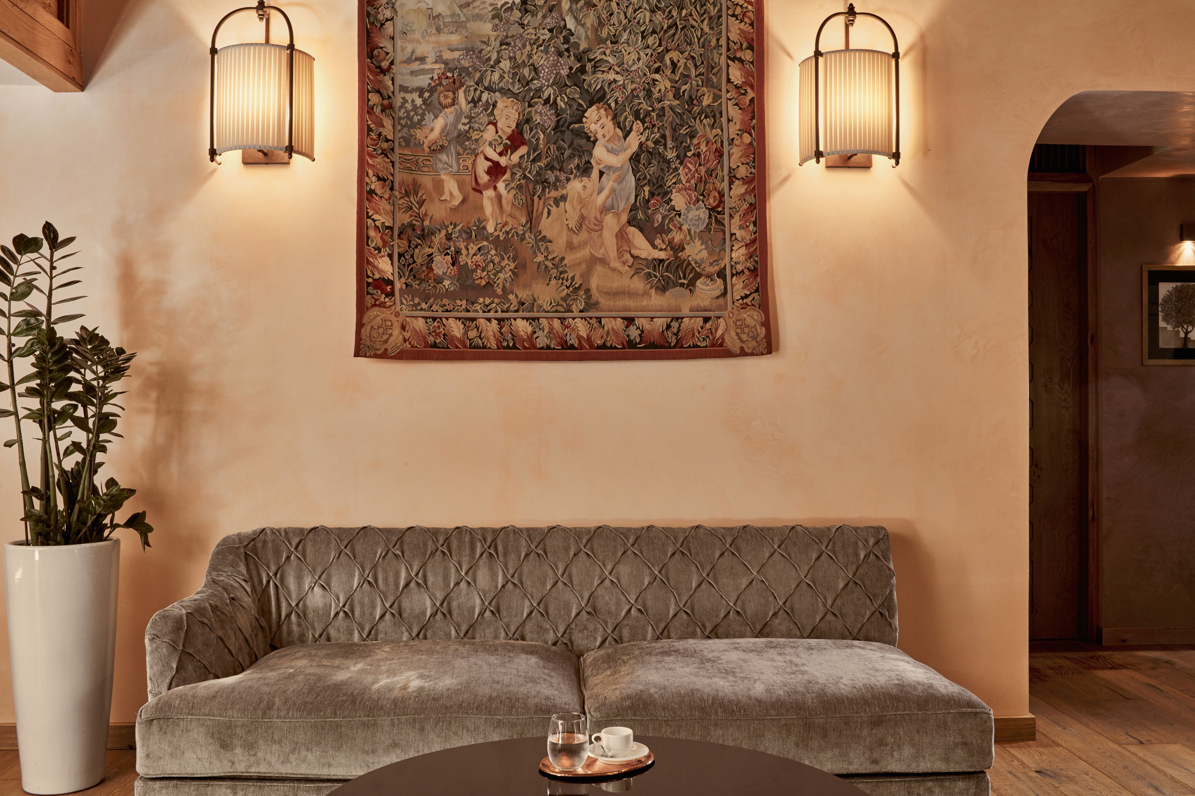 A plush grey sofa with a diamond-patterned backrest, a round coffee table with a glass and cup, and a large tapestry with a nature scene.