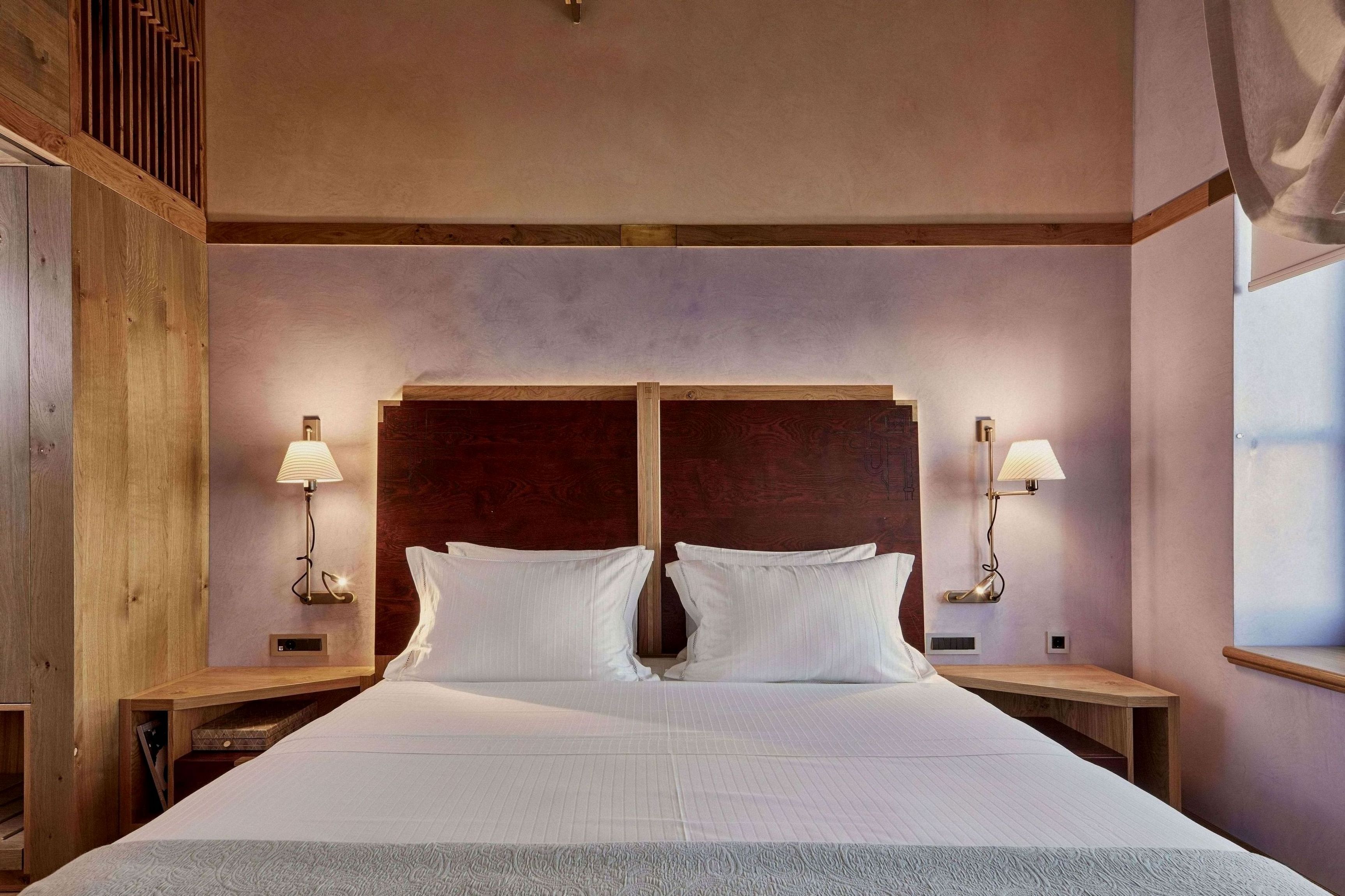 Una cama doble con cabecera de madera, dos mesitas de noche con lámparas y una ventana a la derecha.