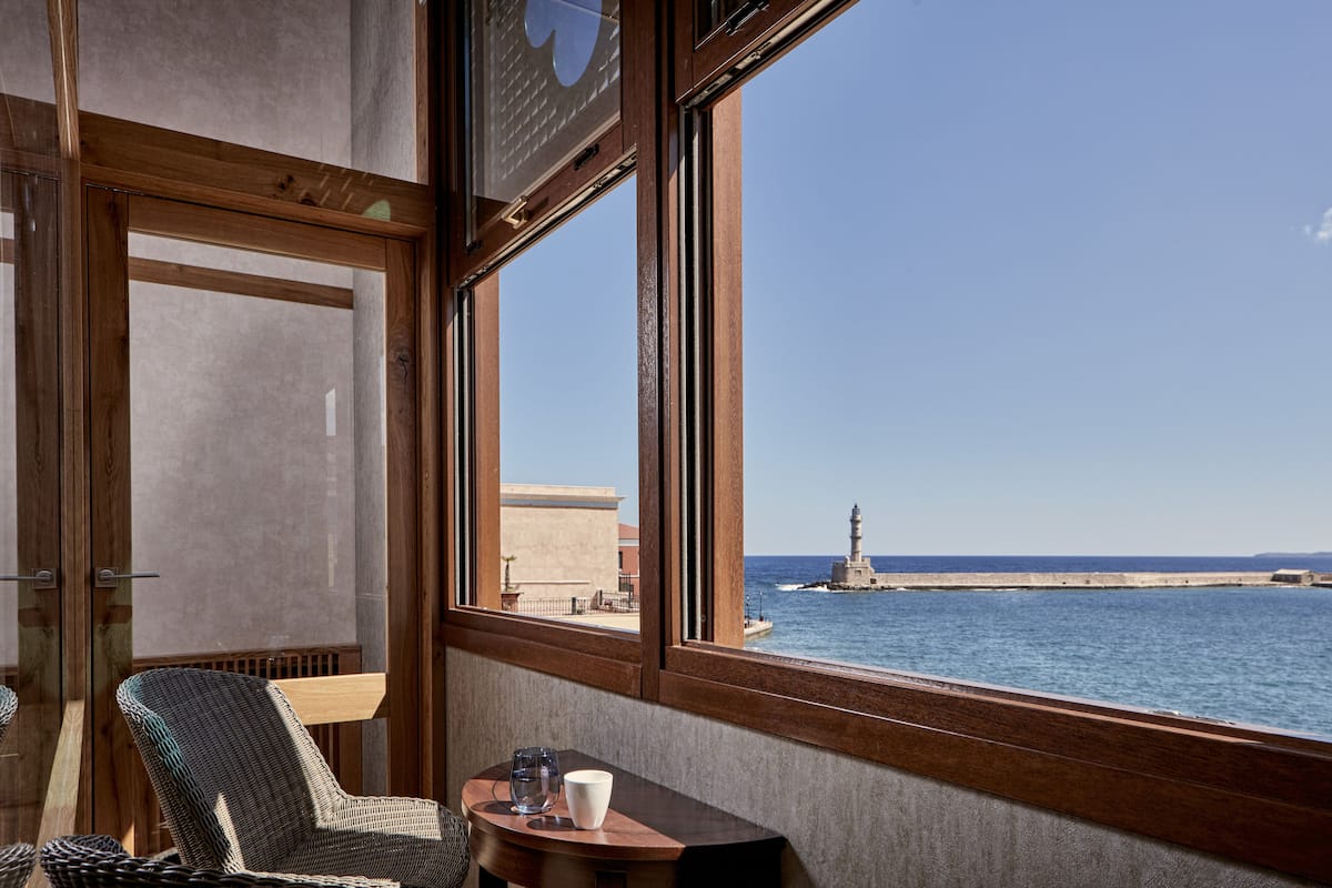 A room with a large window overlooking a body of water and a lighthouse. There is a wooden chair and a small table with a cup and glass.