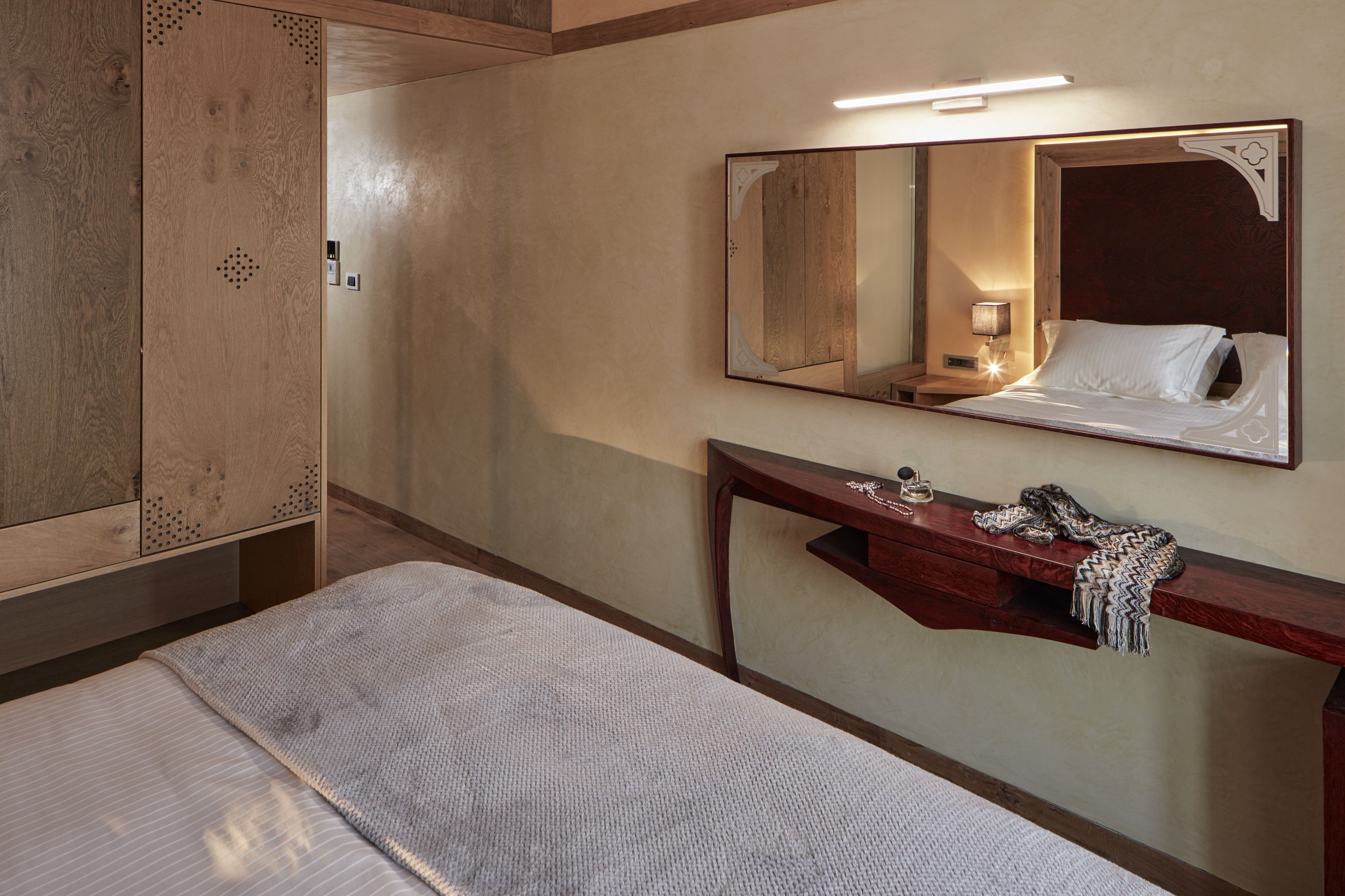 A hotel room with a bed, a wooden wardrobe, a vanity with a mirror, and a bedside table with a lamp and decorative items.