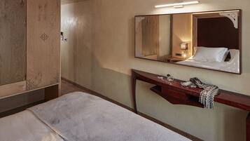 A hotel room with a bed, a wooden wardrobe, a vanity with a mirror, and a bedside table with a lamp and decorative items.