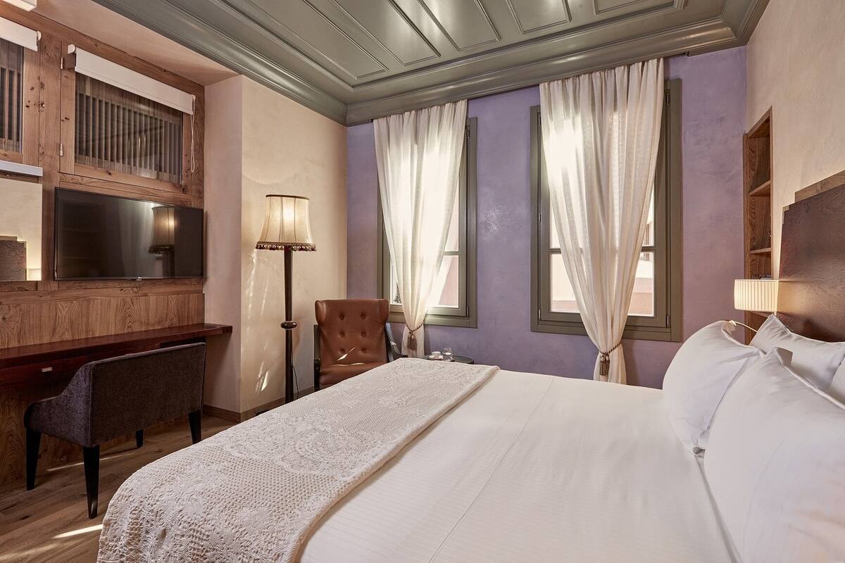 A hotel room with a large bed, a desk, a chair, a television, and two windows with curtains.