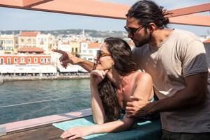 Una pareja en un balcón con vista al frente marítimo y edificios al fondo.