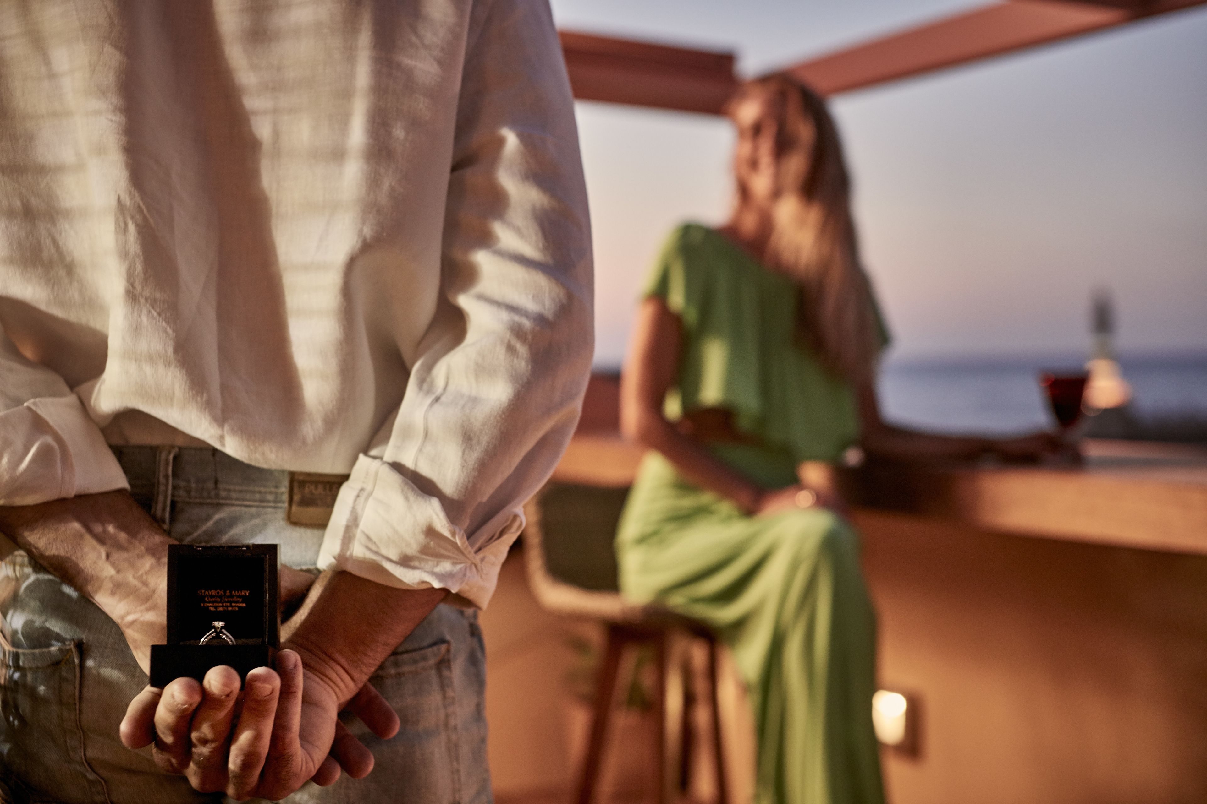 A man holding a ring box, a woman in a green dress seated at a bar, and a view of the sea in the background.