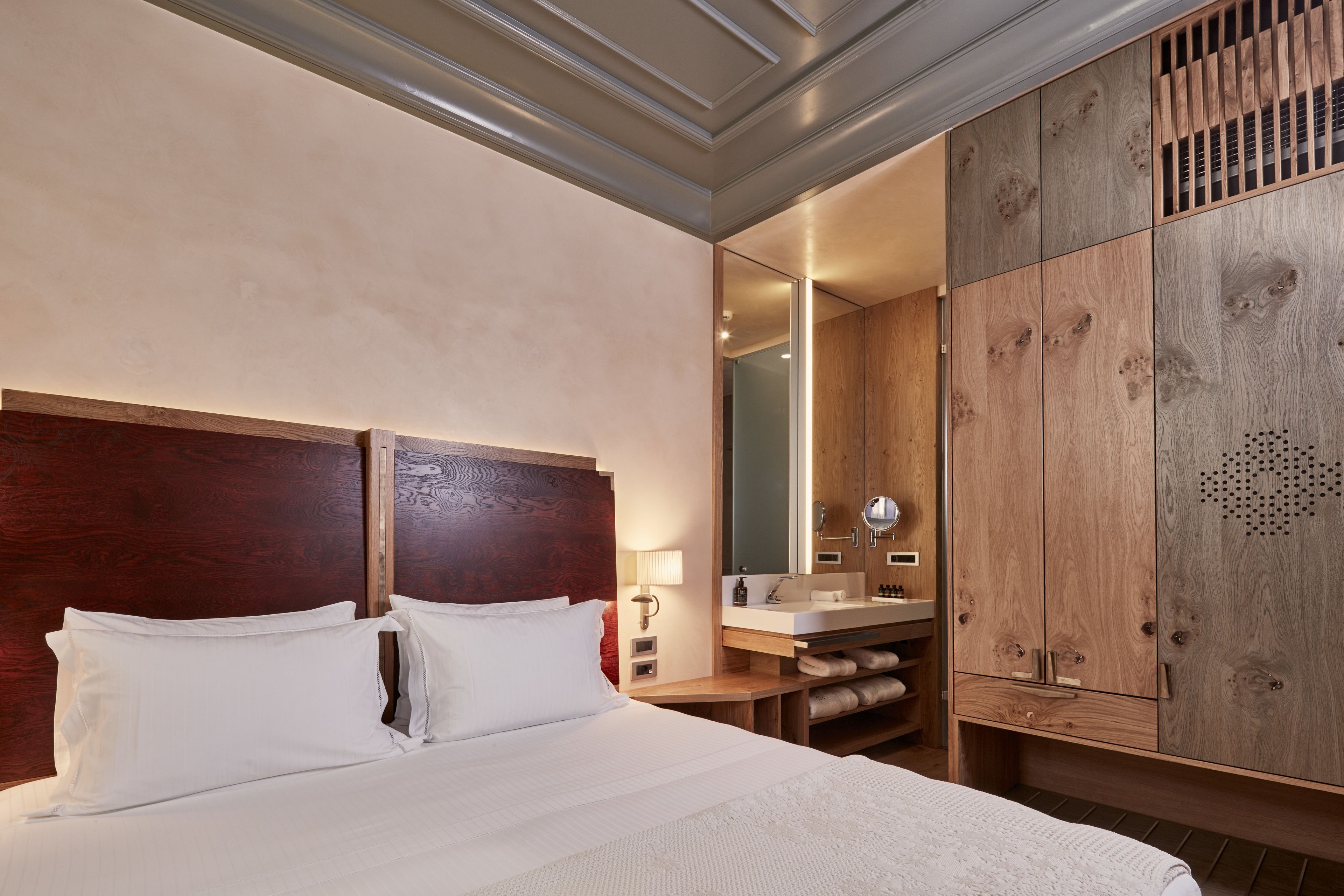 Quarto de hotel com uma cama grande, cabeceira de madeira, guarda-roupa de madeira, banheiro com pia e espelho, e uma pequena mesa de cabeceira com abajur.