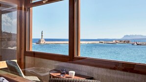 Una habitación con amplios ventanales que dan a una vista costera, con un faro y un puerto con barcos.