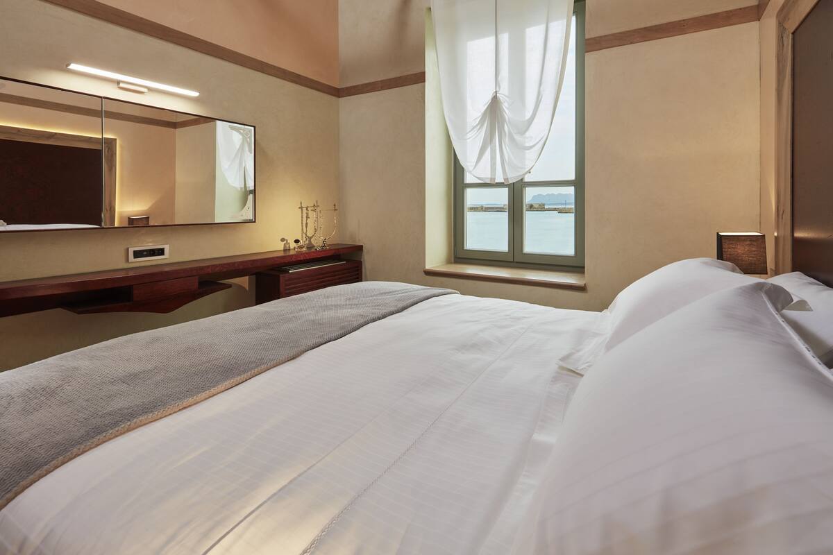 A hotel room with a large bed, a vanity with a mirror, a window with curtains, and a view of the water.