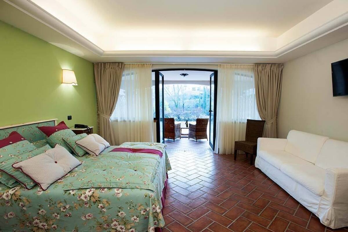 A hotel room with a bed, a white sofa, a TV, and a view through a doorway to an outdoor area with chairs.