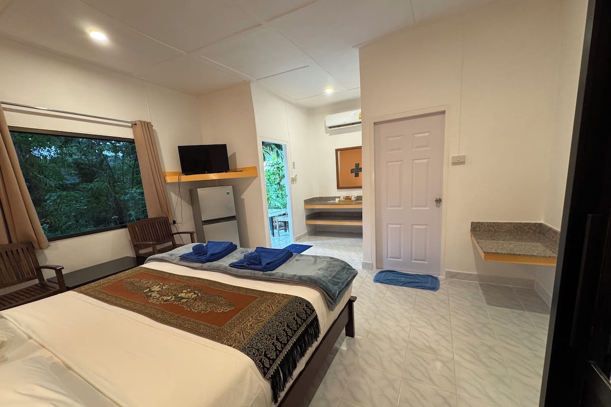 Quarto de hotel moderno com uma cama grande, televisão, frigorífico e varanda com vista para a floresta.