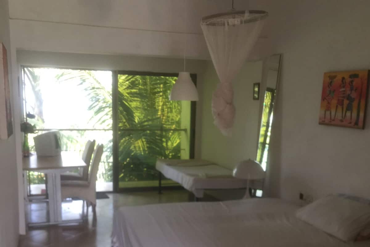 Ein Schlafzimmer mit Bett, Nachttisch, Stuhl und Blick auf einen Balkon mit Pflanzen.